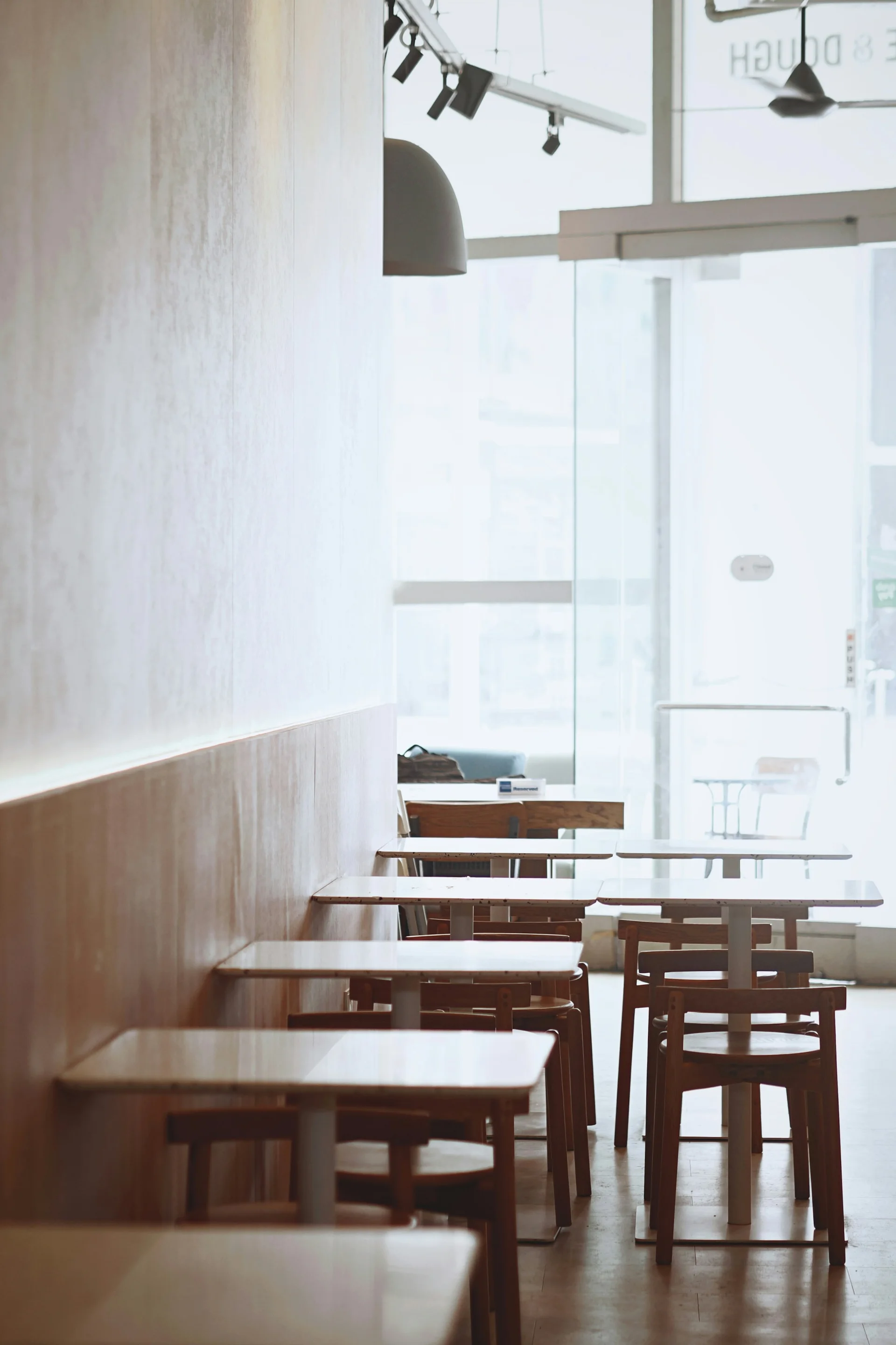 Wooden café tables and chairs repeating along a pale wall beside a sunlit window.